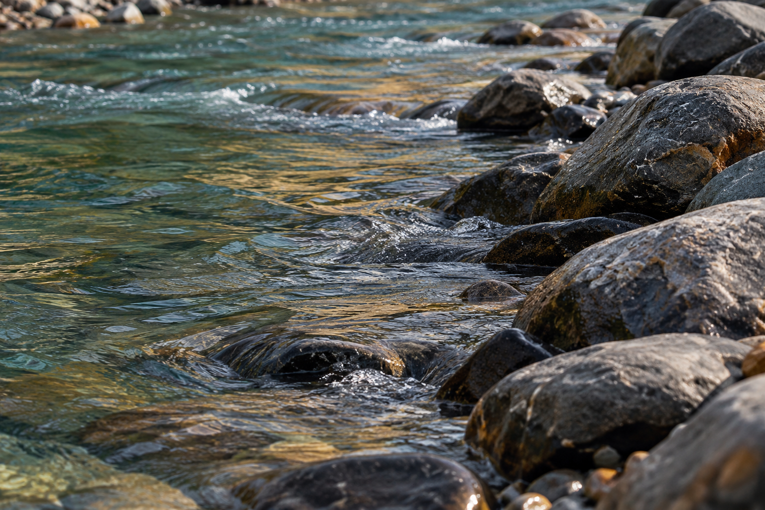 Tight view of moving river water beside dark inland stones in warm afternoon light.